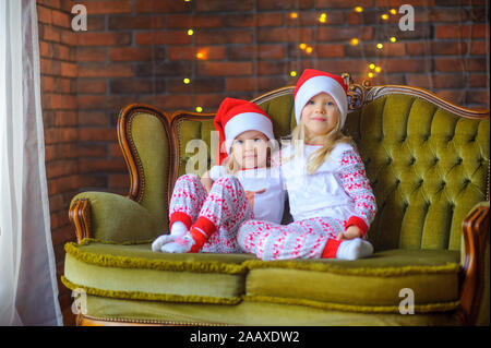 Due graziosi piccole sorelle in un cappello da Babbo Natale sono seduti sul divano in camera. Attesa per le vacanze Foto Stock