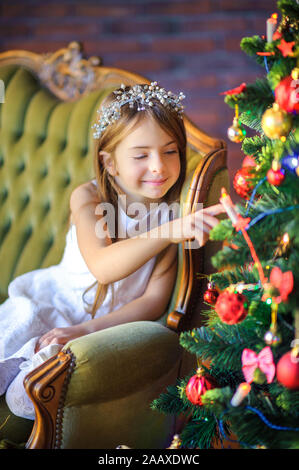 Carino bambina si siede su un divano vicino a un festoso albero di Natale, tocchi e ritiene le decorazioni di Natale. Foto Stock