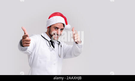 Bel giovane medico in bianco uniforme e Santa Claus hat in piedi in studio su sfondo bianco sorriso e mostrando un dito nella fotocamera studente di medicina Foto Stock