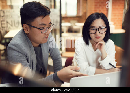 Ritratto di due moderni Asian business persone per discutere progetti di avvio mentre puntando al computer portatile in ufficio Foto Stock