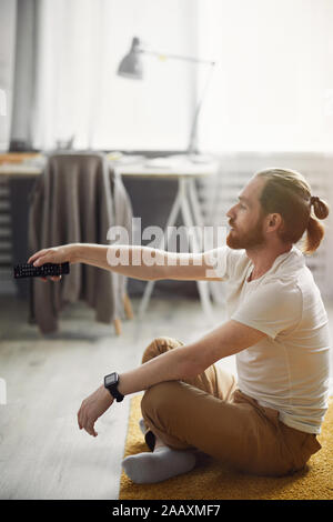 Vista laterale verticale della moderna giovane commutare i canali mentre si guarda la TV di casa seduti sul pavimento Foto Stock