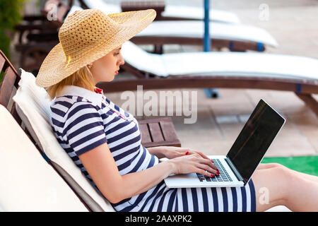 Vista laterale ritratto della bella grave giovane adulto scrittore woman in hat e il vestito è seduta sul letto accogliente con computer portatile sul bordo della piscina e la digitazione di libro. L Foto Stock