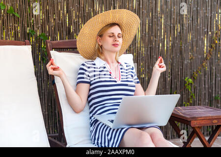 Ritratto di bella calma giovane adulto freelancer donna in hat e il vestito è seduta sul letto accogliente con laptop e avere break, meditando con chiuso Foto Stock