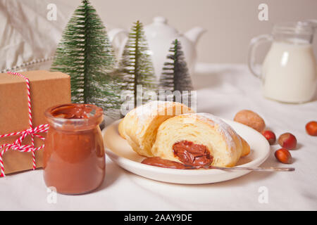 Cornetti freschi bun con cioccolato sulla piastra, una tazza di caffè, un vasetto di latte nelle vicinanze, tre piccolo albero di Natale giocattolo e confezione regalo sul bianco backgro Foto Stock