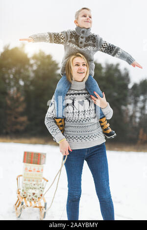 Giovani piuttosto sorridente madre e il suo piccolo figlio seduto su mom sholudres, guardando in alto, godendo di una passeggiata all'aperto con legno decorato slitta in snowy wi Foto Stock