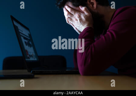 Sottolineato giovane uomo lavora fino a tardi sul laptop, sentendosi frustrato e depresso che copre il volto con le mani a tarda sera Foto Stock