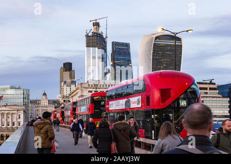 City of London, Londra, Regno Unito - 8 novembre 2018. Coda di autobus rossi come pendolari a piedi attraverso il London Bridge durante le ore di punta con sfondo dei grattacieli Foto Stock