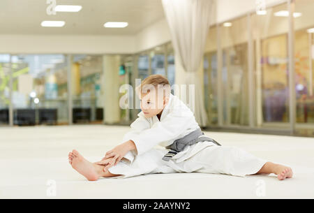 Poco karateka in kimono facendo esercizi di stretching sul pavimento in palestra Foto Stock