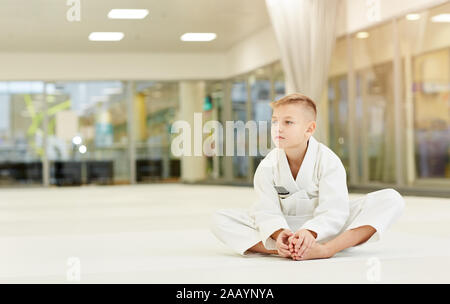 Little Boy in kimono bianco seduto sul pavimento e riposo dopo la formazione sportiva nel karate in palestra Foto Stock