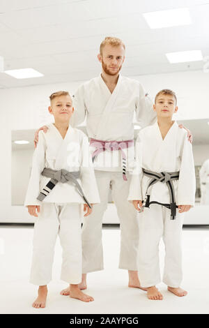 Ritratto di due giovani judoists in kimono in piedi tra il coach e sorridente alla fotocamera in palestra Foto Stock