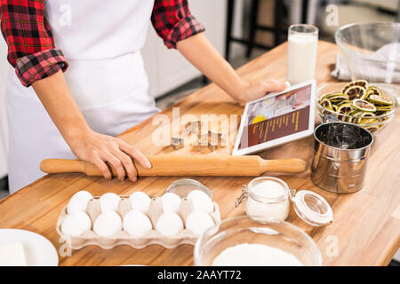 Le mani del giovane massaia holding mattarello e tablet con ricetta mentre in piedi da tavolo in legno e andare a cucinare pasta Foto Stock