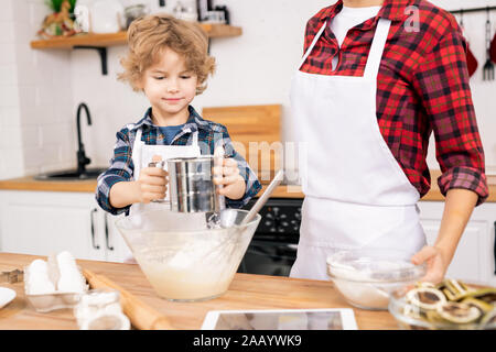 Carino piccolo ragazzo setacciare la farina permanente, mentre dalla tabella accanto alla sua mamma e preparare l'impasto per dolci fatti in casa Foto Stock