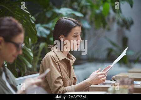 Vista laterale Ritratto di giovane imprenditrice la lettura dei documenti mentre si lavora in outdoor cafe, spazio di copia Foto Stock