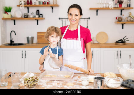 Carino il ragazzo sorridente con il buratto e di sua madre con il mattarello in piedi accanto al tavolo della cucina mentre rendendo i cookie insieme Foto Stock