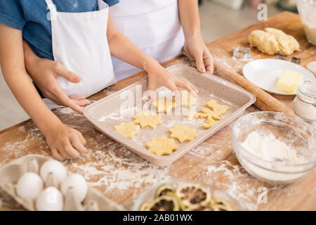 La mano del giovane mettendo materie biscotti sul vassoio mentre aiuta la mamma con la pasta e i cookie da tavolo da cucina Foto Stock