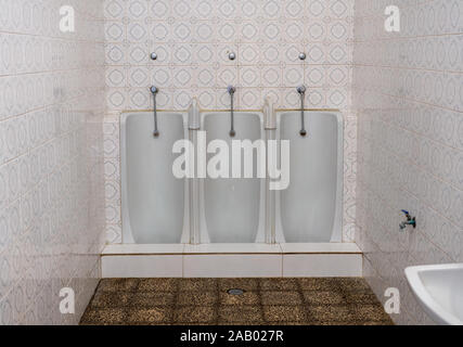 Set di tre fianco a fianco per tutta la lunghezza del Mens di orinatoi in porcellana contro una parete piastrellata in piccole toilette Foto Stock