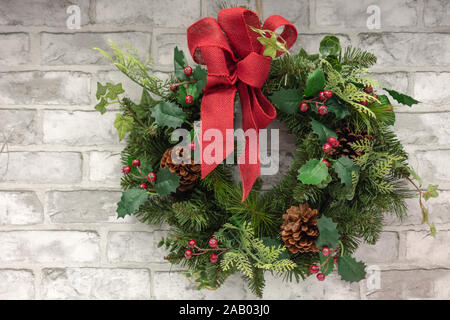 Decorazione di Natale corona con red holly bacche, abete coni e nastro rosso sulla prua di mattoni grigi di sfondo a parete. Foto Stock