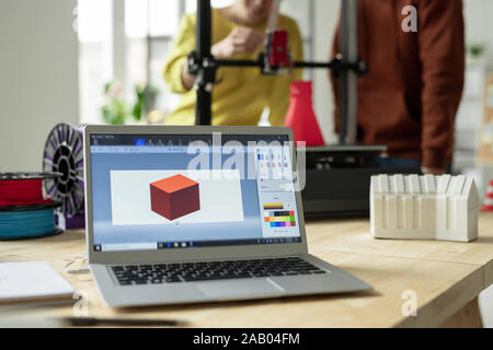 Luogo di lavoro creativo di ingegnere progettista o con il computer portatile e le foto del nuovo modello 3d sul display sullo sfondo di due dipendenti Foto Stock