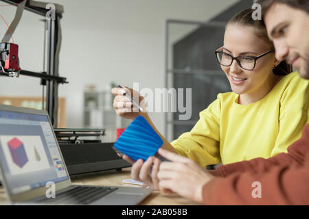 Due giovani progettisti contemporanei discutendo blu oggetto in plastica nella parte anteriore del computer portatile dopo la stampa su stampante 3D Foto Stock