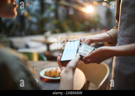 Close up della giovane donna di pagare tramite NFC in cafe, concentrarsi sulle mani femminili holding smartphone con schermo vuoto al terminale bancario, spazio di copia Foto Stock