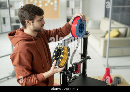 Giovane uomo creativo cambiando lo stelo con filamento permanente, mentre da 3d stampante prima della stampa di oggetti di vari colori Foto Stock