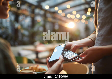 Primo piano della giovane donna di pagare tramite NFC in cafe, concentrarsi sulle mani femminili holding smartphone con schermo vuoto al terminale bancario, spazio di copia Foto Stock