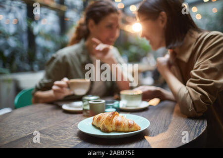 Ritratto di due giovani donne spettegolare mentre vi gustate il pranzo insieme in cafe, focus su croissant fresco in primo piano, spazio di copia Foto Stock