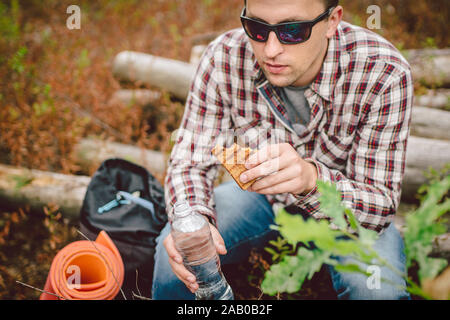 Fast food snack a tema sulla natura. Uomo caucasico mangiare un panino e acqua potabile da una tavolozza bottiglia nella foresta. Tourist arrestato per pranzo in Foto Stock