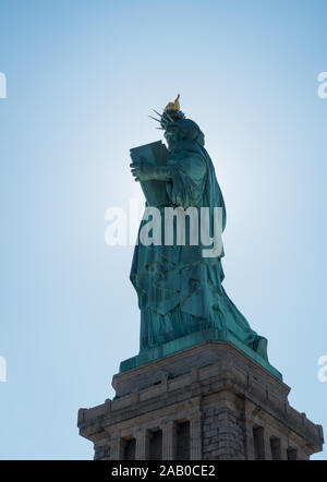 Torna shot dalla statua della libertà con il sole che tramonta dietro la statua e bordi incandescente. Torcia, piedistallo e cielo blu visibile. Riprese in condizioni di luce diurna. Foto Stock