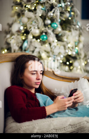 Giovane donna controllo fuori i suoi messaggi di testo sul telefono cellulare come ella liyng divano nella parte anteriore dell'albero di Natale a casa. Foto Stock