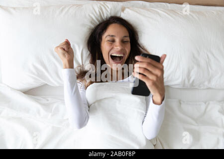 La donna si trova nel letto dello smartphone di contenimento urlare di gioia Foto Stock