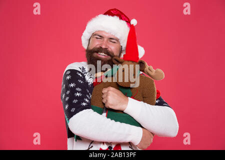 Abbraccio e l'amore. Acquistare regali. Inverno vendita. Festa d'inverno. La renna. L'uomo santa hat tenere regalo di Natale sfondo rosso. Natale concetto prep. Tanga faccia barbuta inverno maglione. Vacanze invernali. Giocattolo morbido. Foto Stock