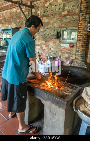 In Cai Be, Mekong Deltal, Vietnam - Marzo 13, 2019: uomo in maglietta blu di ramoscelli mette a fuoco per illuminare la sua black IVA al pop chicchi di riso contro brown bric Foto Stock