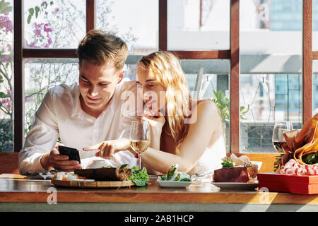 Coppia giovane guardare qualcosa di interessante sullo smartphone quando avente una cena romantica nel ristorante Foto Stock