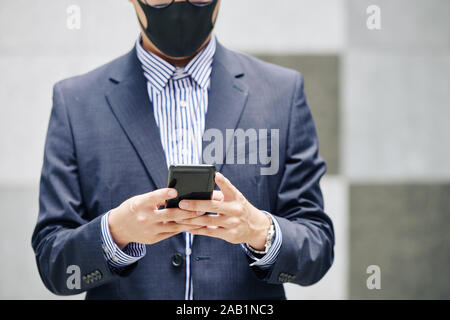 Immagine ritagliata di imprenditore in maschera protettiva texting collega o controllare i social media Foto Stock