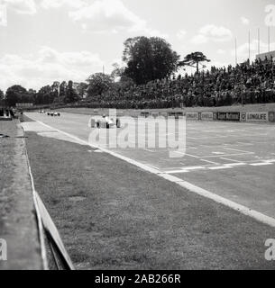 Anni sessanta, storico motor racing al Crystal Palace il circuito di gara in Crystal Palace Park nel Sud di Londra, London, England, Regno Unito, una vettura scende per la finitura guardato dritto da una folla di spettatori. Foto Stock