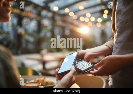 Primo piano della giovane donna di pagare tramite NFC in cafe, concentrarsi sulle mani femminili holding smartphone con schermo vuoto al terminale bancario, spazio di copia Foto Stock