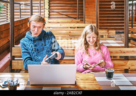 Giovane ragazzo gioca a un computer portatile in un cafe. La ragazza nelle vicinanze è la lettura di un libro. concetto di usura Foto Stock