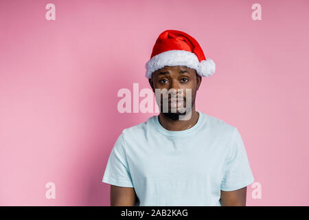 Bello grave barbuto americano africano giovane uomo che indossa red Santa hat, bianco di t-shirt, guardando la fotocamera con calma, fiducia espressioni, isolato Foto Stock