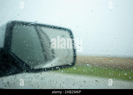 La guida su una strada una piovosa giornata di primavera. Le gocce di pioggia sul vetro dell'auto e sul parafango specchio, cielo nuvoloso, campagna di sfocatura Foto Stock
