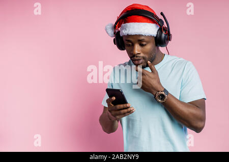 Sorpreso uomo africano con setola, indossare un cappello da Babbo Natale, bianco di t-shirt, ascoltando musica in cuffia, tenendo il telefono cellulare, guardando lo schermo con amaz Foto Stock