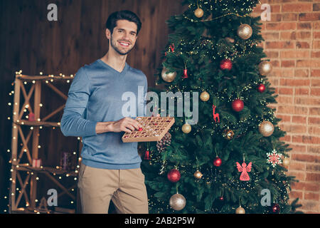 Foto di allegro positivo uomo carino in blu maglione beige Pantaloni Pantaloni sorridente finitura toothily decorare albero di natale Foto Stock