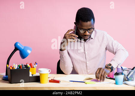 Pragmatico barba lunga african american uomo che indossa gli occhiali, camicia a righe, parlando con il partner aziendale sullo smartphone, seduti alla scrivania circondata wit Foto Stock