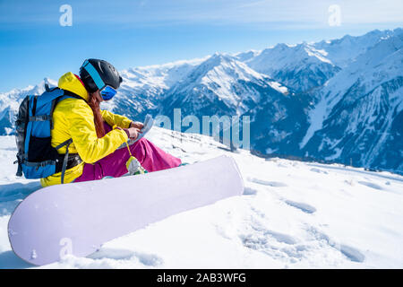 Immagine di snowboarder ragazza nel casco con mappa nelle sue mani seduto sul pendio della montagna in inverno Foto Stock