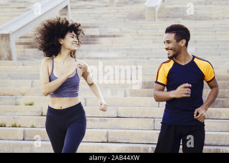 Donna sorridente e uomo felice in esecuzione al piano di sotto sulla città di scale, fitness, urban allenamento sportivo e di uno stile di vita sano concetto Foto Stock