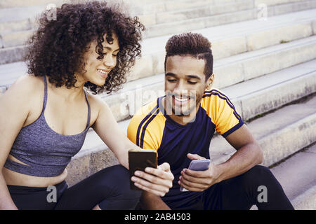 Felice atletica donna latina mostra le app di sport nel suo telefono mobile ad un nero uomo sportivo sulle scale durante il resto dell'allenamento, tecnologia in sp Foto Stock