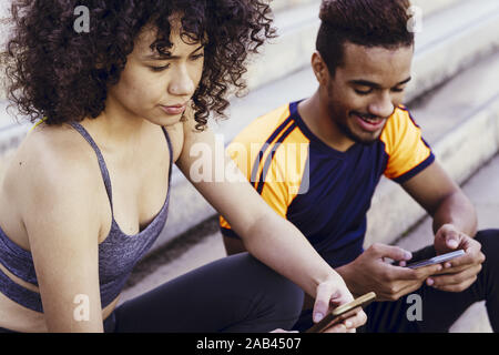 Ritratto di un latino atletica donna e uomo nero utilizzando i loro telefoni cellulari sulle scale durante il resto dell'allenamento, la tecnologia nello sport e sano Foto Stock