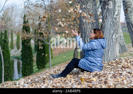 La donna di mezza età è giocare con le foglie di autunno in autunno Foto Stock