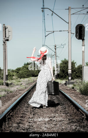 Ragazza giovane con la vecchia valigia su binari ferroviari. Una bella ragazza sta camminando con una valigia. La vista dal retro. Un adolescente in una tartina hat con Foto Stock