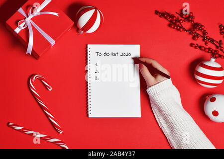 Mano femminile in maglia maglione iscritto Lista da fare sul notebook su sfondo rosso. Nuovo anno 2020 il concetto di pianificazione. Foto Stock